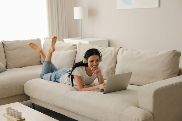 Woman with laptop and headphones lying on sofa at home - Fotó, kép