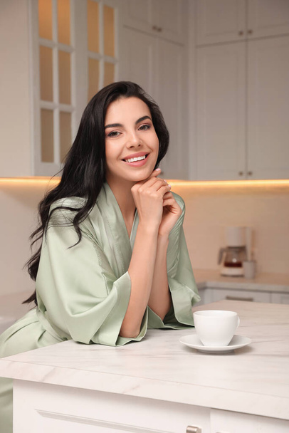 Pretty young woman wearing beautiful silk robe with cup of coffee in kitchen - Foto, Imagen