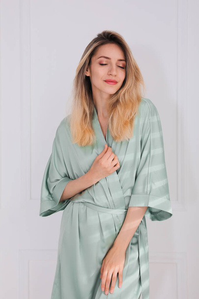 Pretty young woman in beautiful light green silk robe near white wall indoors - Фото, изображение