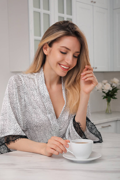 Pretty young woman in beautiful silk robe with cup of coffee at kitchen table - 写真・画像