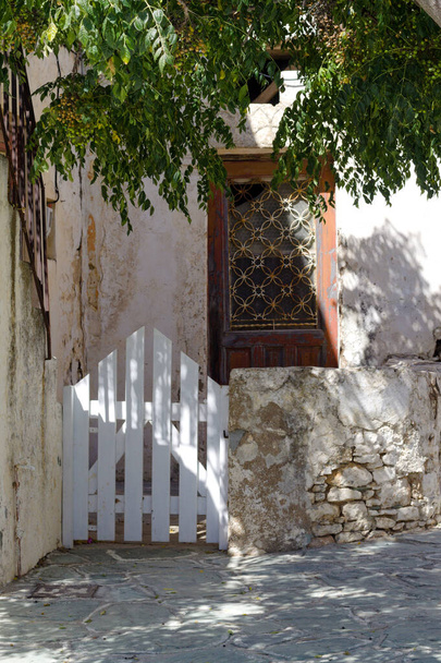 Φολέγανδρος, Ελλάδα. Σπίτι στο χωριό στην παλιά πόλη, το Κάστρο. Γοητευτική, απλή παραδοσιακή ελληνική νησιώτικη σκηνή. Λεπτομέρεια πρόσοψης ακινήτου με πύλη και πόρτα σε σκιασμένη αυλή. Κάθετη βολή - αντιγραφή χώρου. - Φωτογραφία, εικόνα