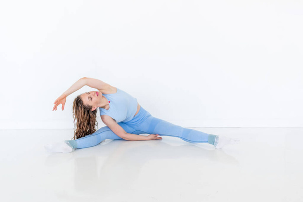 Young diverse woman practices yoga, doing body stretching exercise. Sitting on the floor. White studio loft. Sports and wellness concept. Copy space - Фото, зображення