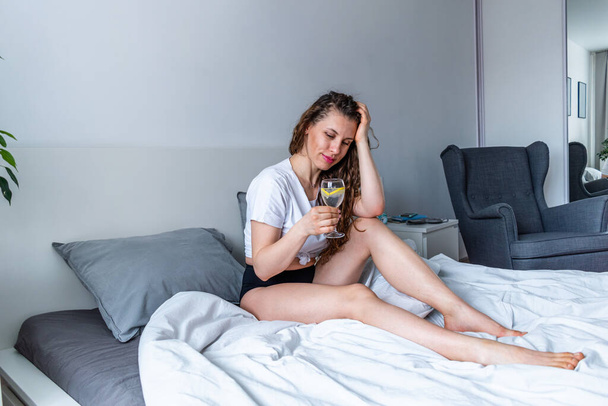 Young woman ginger hair sitting the bed in bedroom and drinks water with lemon. Fresh morning. Wellness and detox concept. Copy space - 写真・画像