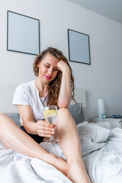 Young woman ginger hair sitting the bed in bedroom and drinks water with lemon. Fresh morning. Wellness and detox concept. Copy space - Фото, зображення
