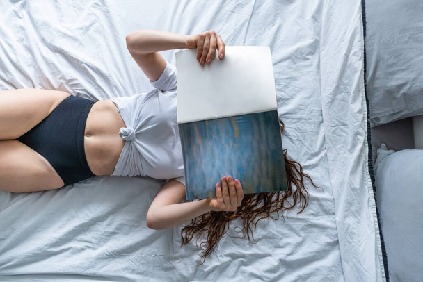 Young woman lies on bed and reads a magazine. Concept no photo face. Beautiful fit athletic body - Fotó, kép