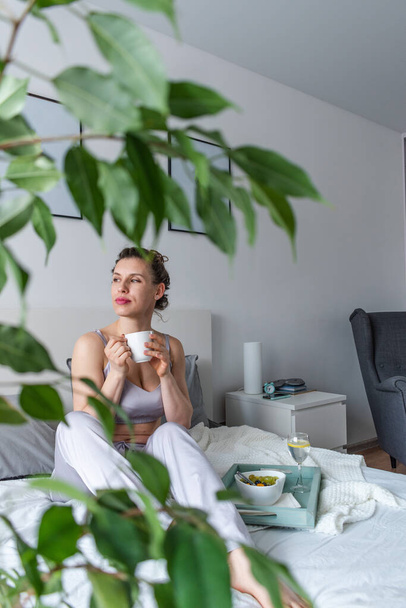Young woman in silk pajamas has healthy breakfast while lying on bed in bedroom. Home wellbeing concept. Emotional health of a young woman - Foto, immagini