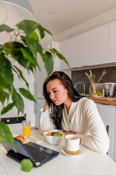 Young woman in comfortable white robe works on laptop while sitting at table the kitchen. Work at home for breakfast. Business woman maintains social distance, works remotely. New normal concept - Photo, image