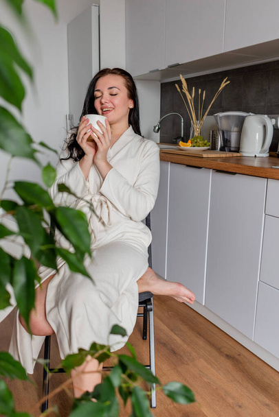Young woman with black hair in white robe sits on chair the kitchen. Hands hold white mug with aromatic coffee. Wellness and healthy food concept - Фото, изображение