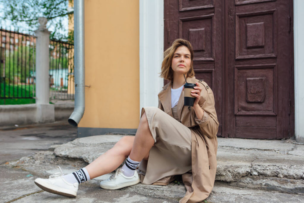 Young millennial woman with wild hair dressed in an autumn coat sitting near the door of an old building with coffee to go. - Фото, изображение
