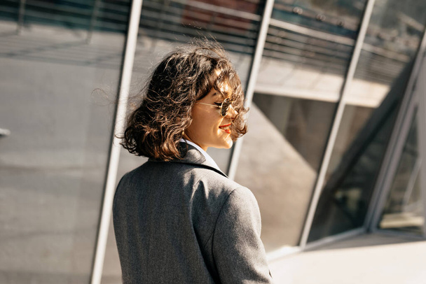 Lifestyle portret van vrij stijlvol meisje met korte krullend kapsel wandelen in de stad in zonlicht. Gelukkige jonge vrouw met een zonnebril en een jasje glimlacht buiten. Hoge kwaliteit foto - Foto, afbeelding