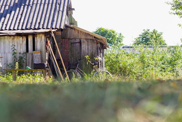Antiguo granero abandonado en ruinas desenfocado en el patio sobre un fondo verde. Varios utensilios cerca de él. Una vieja bicicleta en la puerta. Una vida rural sencilla. fuera de foco. - Foto, imagen