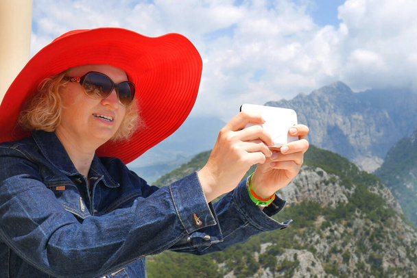 Jolie jeune femme en chapeau rouge utilisant son téléphone portable pour faire des photos dans les montagnes - Photo, image