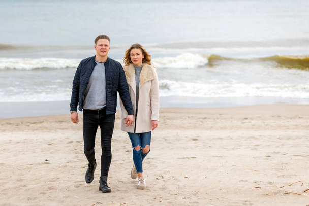 Atractiva pareja joven caminando a lo largo de la orilla de una playa de arena, en unas vacaciones románticas de primavera, al aire libre.Pareja dando un paseo cogidos de la mano en la playa.Pareja disfrutando de un tiempo amoroso juntos. - Foto, Imagen