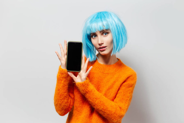 Portrait of young beautiful girl with blue hair holding smartphone in hands on background of light grey; wearing orange sweater. - Photo, image