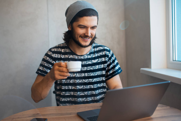 Портрет молодого щасливого чоловіка, який п'є каву під час роботи на ноутбуці
. - Фото, зображення