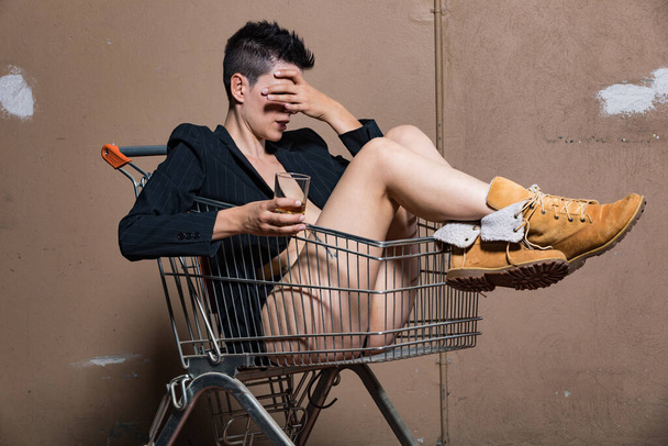 Woman sitting in a shopping cart is hiding the visono with a glass of cognac in her hand. She is half naked - Photo, image