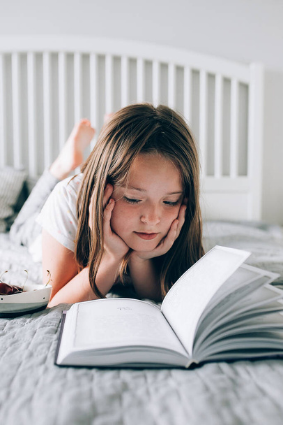 8-9 yaşlarında güzel bir kız yatağa uzanıp kitap okuyor. Rahat, rahat ev konsepti. Yaşam biçimi - Fotoğraf, Görsel