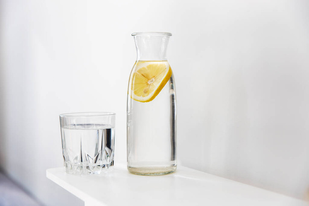 A refreshing glass of water, a bottle with a slice of lemon inside is on the dresser in the summer heat - Fotoğraf, Görsel