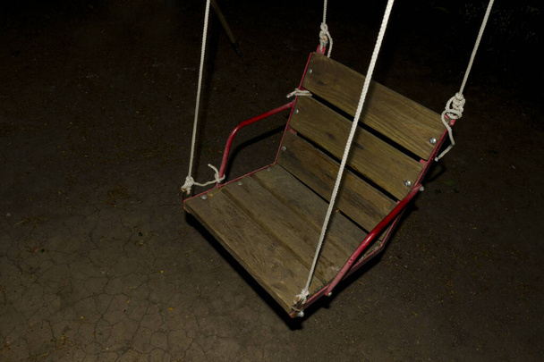 Playground at night, spooky photo. Backlit wooden swing. Isolation, loneliness and sadness concept.A swing at night. - Photo, Image