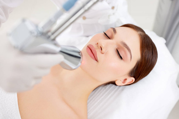 Cosmetólogo profesional que utiliza equipos láser para el tratamiento del cuidado de la piel en la cara del cliente femenino en la clínica de belleza moderna - Foto, imagen