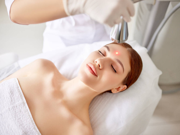 Cosmetólogo profesional que utiliza equipos láser para el tratamiento del cuidado de la piel en la cara del cliente femenino en la clínica de belleza moderna - Foto, Imagen