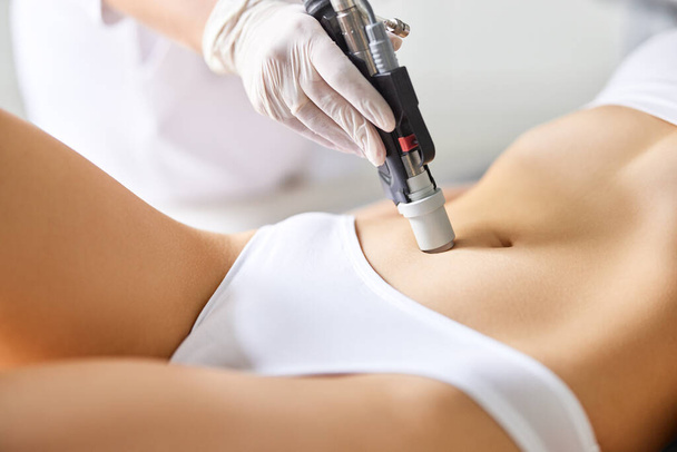 Περικοπή ανώνυμη γυναίκα ασθενή που λαμβάνουν λέιζερ διαδικασία φροντίδας του δέρματος στο στομάχι κατά τη διάρκεια του διορισμού σε επαγγελματική κλινική ομορφιάς - Φωτογραφία, εικόνα