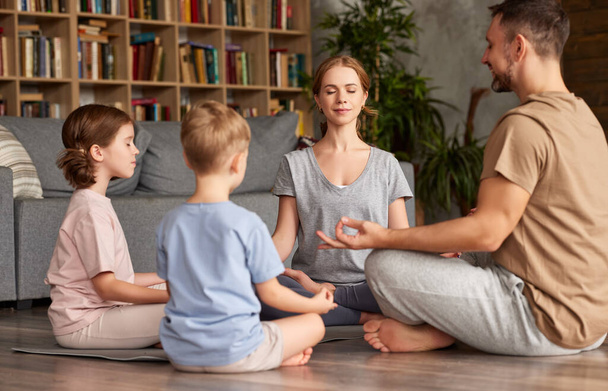 Jeunes parents méditant avec des enfants à la maison. beau père de famille, mère et deux enfants avec les yeux fermés pratiquant le yoga assis en cercle dans la pose du lotus - Photo, image