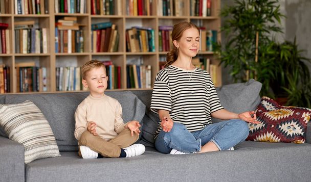 Junge Mutter und Sohn in Freizeitkleidung meditieren mit geschlossenen Augen, während sie im Lotus sitzen und gemeinsam auf dem Sofa im Wohnzimmer posieren. Ruhige schöne Familie praktiziert Yoga während der Quarantäne - Foto, Bild