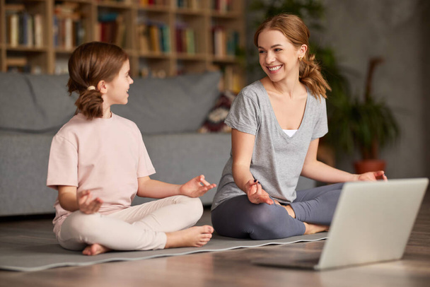 Junge schöne Mutter in Freizeitkleidung lehrt kleine Tochter, wie man meditiert, während man zusammen in Lotus-Pose auf dem Boden im Wohnzimmer zu Hause sitzt, glückliche Familie macht Yoga-Übungen online mit Laptop - Foto, Bild