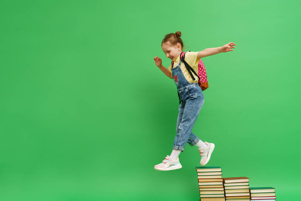 happy little girl of 5 years old in loose clothes with a full-length backpack climbs the steps of books and jumps down on a green background. desire to study. Successful acquisition of knowledge. - Foto, afbeelding
