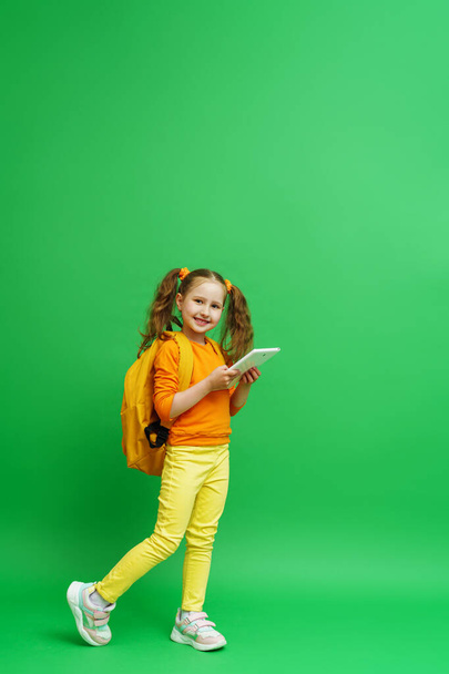 Електронне навчання. мила школярка з двома хвостами з рюкзаком і планшетним комп'ютером в руках стоїть на зеленому тлі, дитина посміхається і дивиться в рамку. Рекомендація віддаленої онлайн школи
. - Фото, зображення