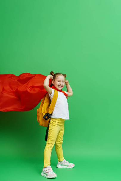 happy little schoolgirl in superhero costume with a red cape and a backpack shows off her muscles. confident, strong and successful little girl smiles and poses on a green background. desire to study. - Foto, Bild