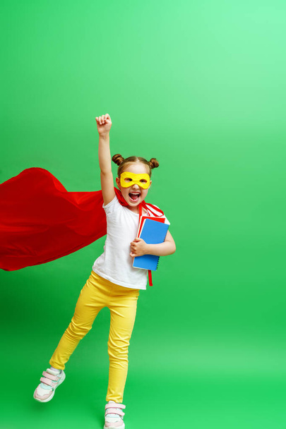 happy smiling schoolgirl in a red cape and a superhero mask and with a school book in her hands,runs and jumps on the green background of the studio. The child is ready for successful learning - Foto, immagini