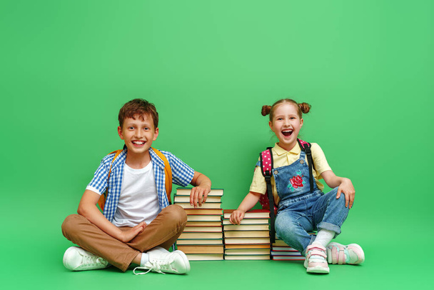 Two happy schoolchildren in loose clothes sit on stacks of school textbooks and reading books, and smile cheerfully against a green background. The concept of children's education. Back to school - Valokuva, kuva
