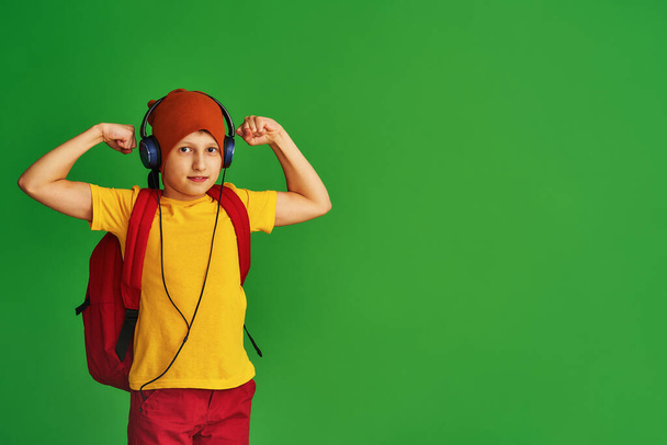 serious Caucasian schoolboy in casual clothes, hat, headphones and school backpack, standing on green background. boy shows his strength, biceps. Return to school. concept Power of education - Zdjęcie, obraz