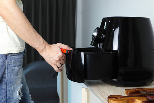 Right hand of man holding the tray of the black deep air fryer or oil free fryer appliance which is on the wooden table in the white kitchen ( air fryer ) during lunch with family members - Foto, immagini