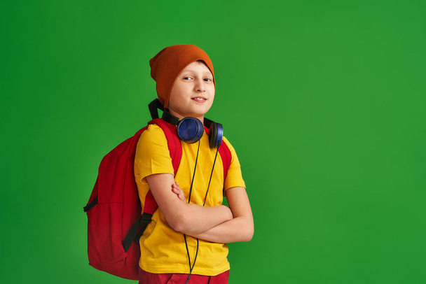 serious Caucasian schoolboy in casual clothes, hat, headphones and school backpack, standing on green background. boy shows his strength, biceps. Return to school. concept Power of education - Foto, imagen