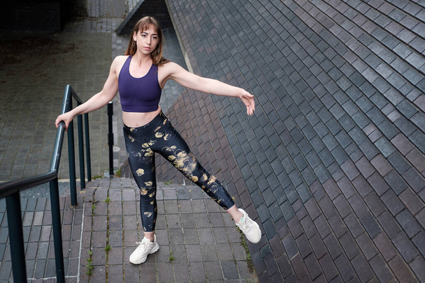Mladá fitness žena drží zábradlí a dělá baletní pozice. Má na sobě pevné aktivní oblečení a je v městském prostředí. - Fotografie, Obrázek