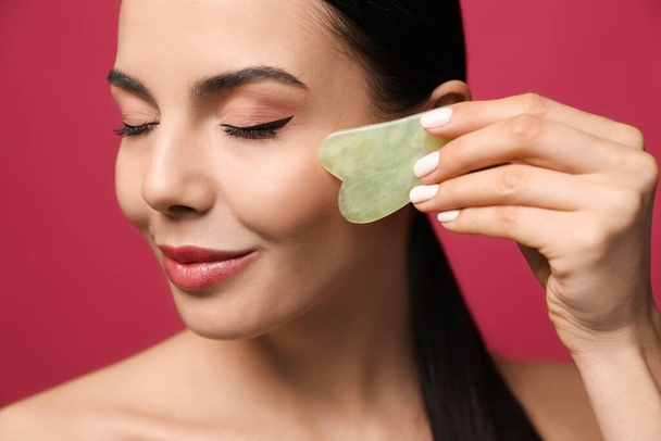 Beautiful young woman doing facial massage with gua sha tool on pink background, closeup - Foto, afbeelding