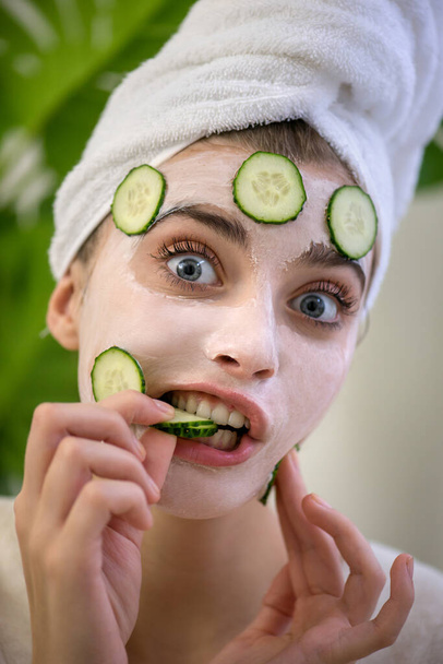 Young woman applies face mask with fresh cucumbers on her face and makes makes a funny face. Beauty model with  perfect fresh skin and long eyelashes cares about her skin at home. Close up, selected focus. - Φωτογραφία, εικόνα