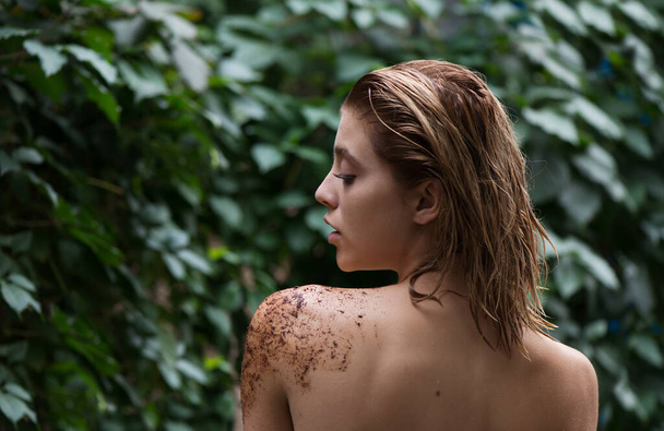 Beautiful young woman uses coffee scrub and peeling on her body front of plant tropical green leaves background.  Model with wet hair care of her face and body. SPA, wellness, bodycare and skincare.  Close up, selective focus. - Фото, зображення