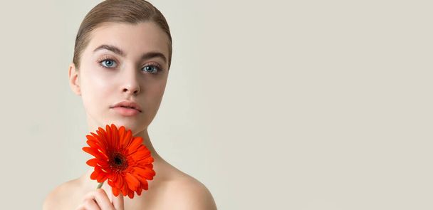 Beautiful young woman with perfect skin and flower. Fashion model face with natural make-up and red  gerbera flower in hand.  SPA, wellness, bodycare and skincare concept. - Фото, зображення