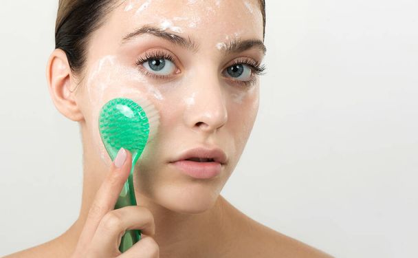 Beautiful young girl cleansing face with cosmetic facial skincare foam, gel or soap by brush. Woman hame SPA procedures. - Photo, image