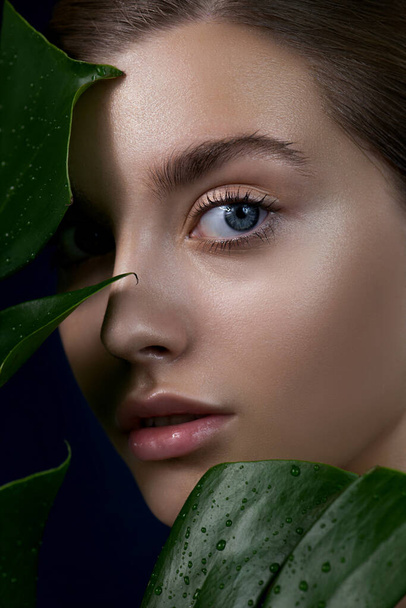 Close-up of beautiful young woman's face with perfect skin through the tropical green leaves. SPA,  wellness and skincare concept.  Close up, selective focus. - Fotoğraf, Görsel