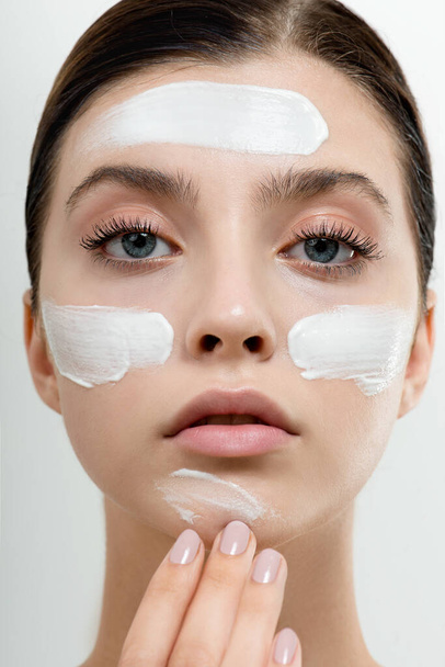Young woman applying face cream or facial mask at her face. Beauty model with perfect fresh skin and long eyelashes cares about her skin at home. Spa and Wellness, Skin Care Concept. Close up, selected focus. - 写真・画像