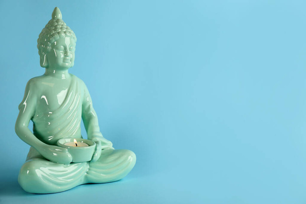 Beautiful ceramic Buddha sculpture with burning candle on light blue background. Space for text - Valokuva, kuva
