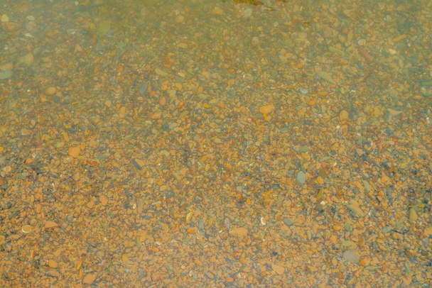 Viele glatte Steine auf dem gelben Sandboden des Gebirgsflusses. Naturhintergrund aus gelbem Lehmboden mit glatten Steinen in Nahaufnahme. Natürliche Textur der Felsbrocken im klaren Wasser des Gebirgsbaches. - Foto, Bild