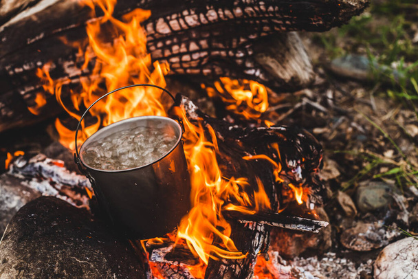Kettle stands on fire. Cooking food at fire in wild. Beautiful big log burns in bonfire close-up. Survival in wild nature. Wonderful flame with caldron. Pot is on flames. Campfire background. - Foto, Imagen