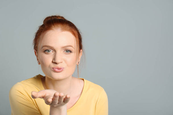 Portrait of young red haired woman blowing kiss on light grey background, space for text - Foto, Imagem