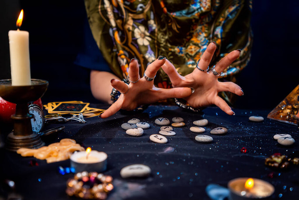 Las manos femeninas de una adivina leen runas. Las cartas del tarot, joyas y velas están sobre la mesa. El concepto de adivinación, astrología y predicción del futuro. - Foto, imagen